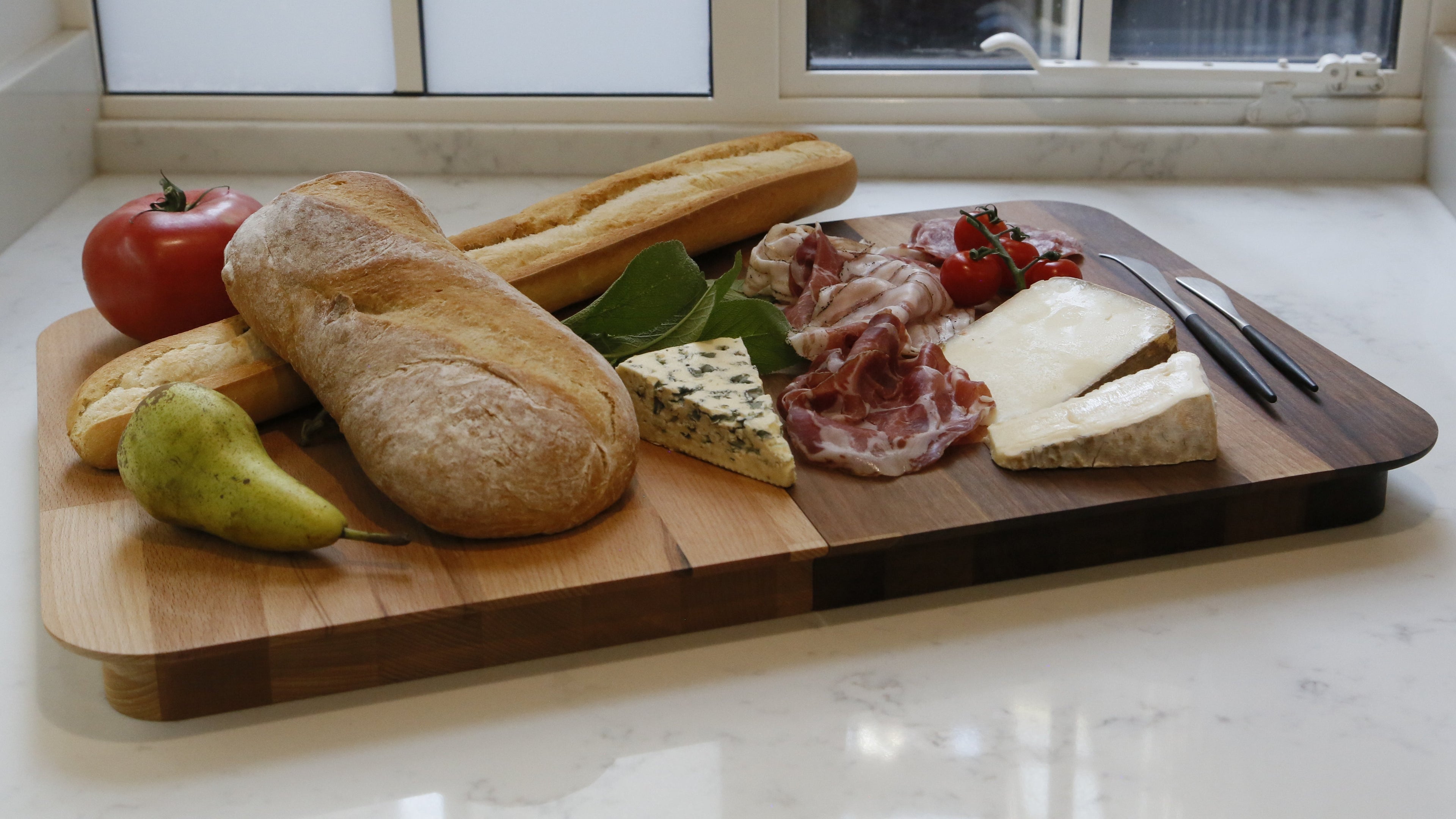 Wooden cutting board with bread, fruits, and meats on a kitchen counter.